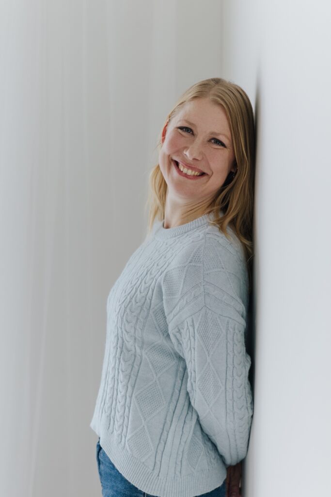 Miina Tauriala, puheterapeutti, hymyilee vaaleassa neuleessa vaaleaa seinää vasten.